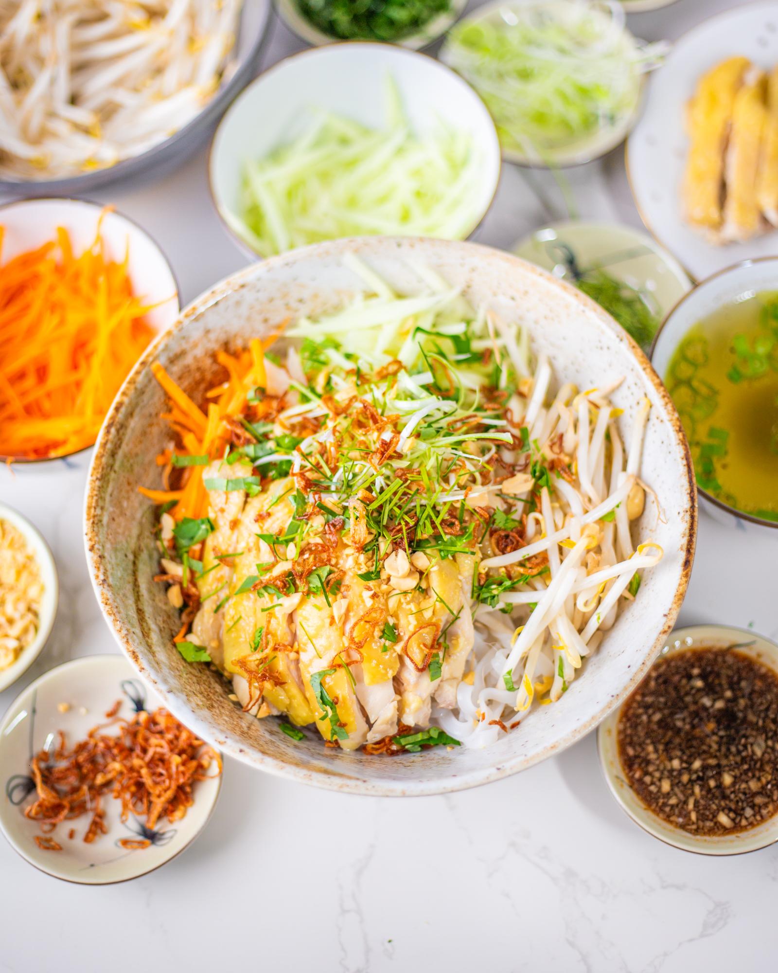 A large white and brown ceramic bowl with shredded carrot, shredded green papaya, bean sprouts, sliced chicken, chopped herbs, peanuts, and fried shallot. It is surrounded by smaller bowls containing the different ingredients.